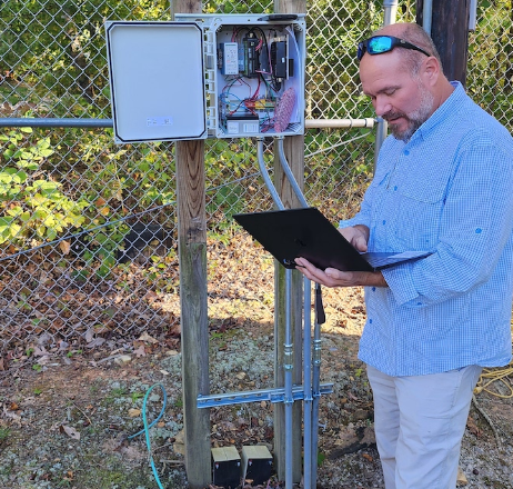 Technician standing at an open control panel by a fence, reviewing data on a tablet in the field.