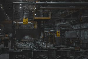 A dimly lit industrial factory interior with machinery, metal pipes, and stacks of materials.