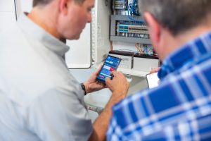 Two people stand in front of an open electrical control panel.